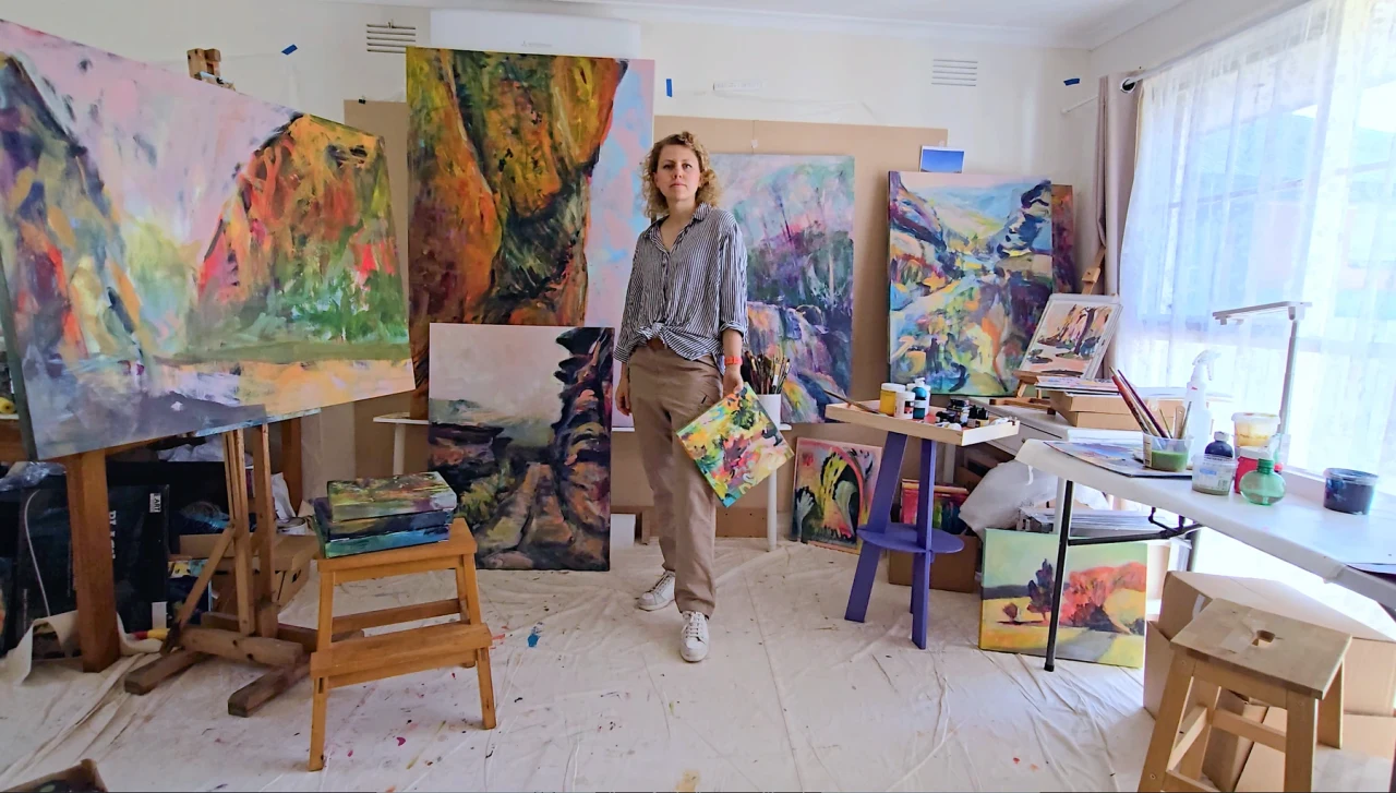 Olga standing in her bright studio, holding a painting with paintings behind her